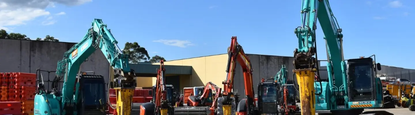 Row of excavators lined up at a construction site, with temporary safety barriers visible in the foreground.