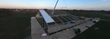 A crane installing a roof panel on a large temporary fabric structure at dusk in an open field